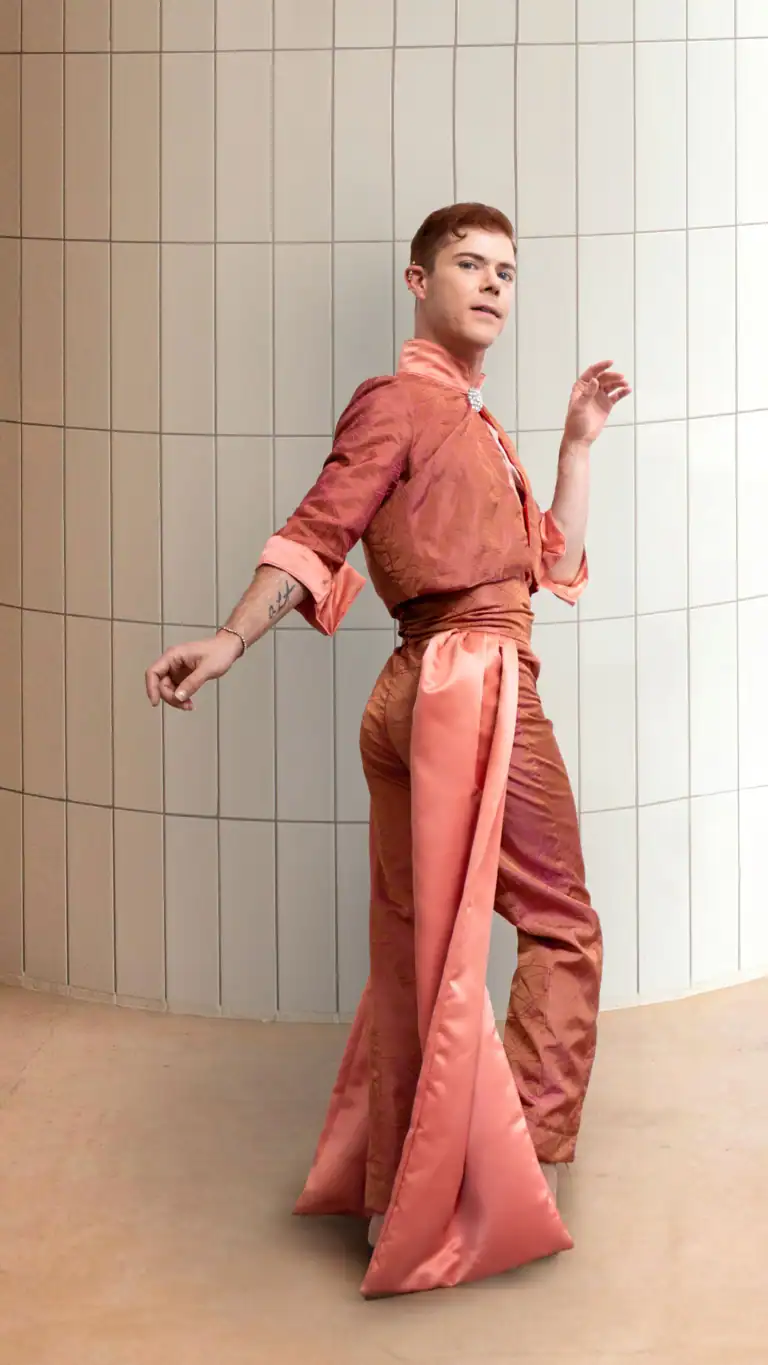 Designs Elijah in a clean white tile room showing his coral flowing suit with feminine details.
