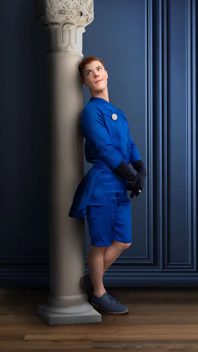 Designs Elijah's feminine fabrics in a unique shorts-style suit as he leans against a column in a blue room.